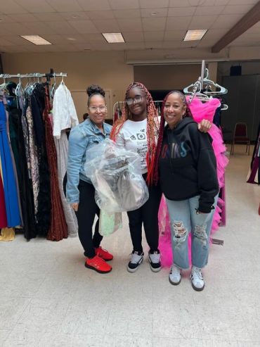Three women smiling among racks of colorful dresses in a room.