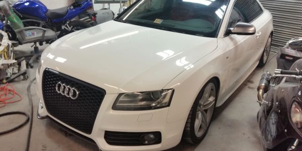 White Audi car parked indoors with motorcycles nearby.