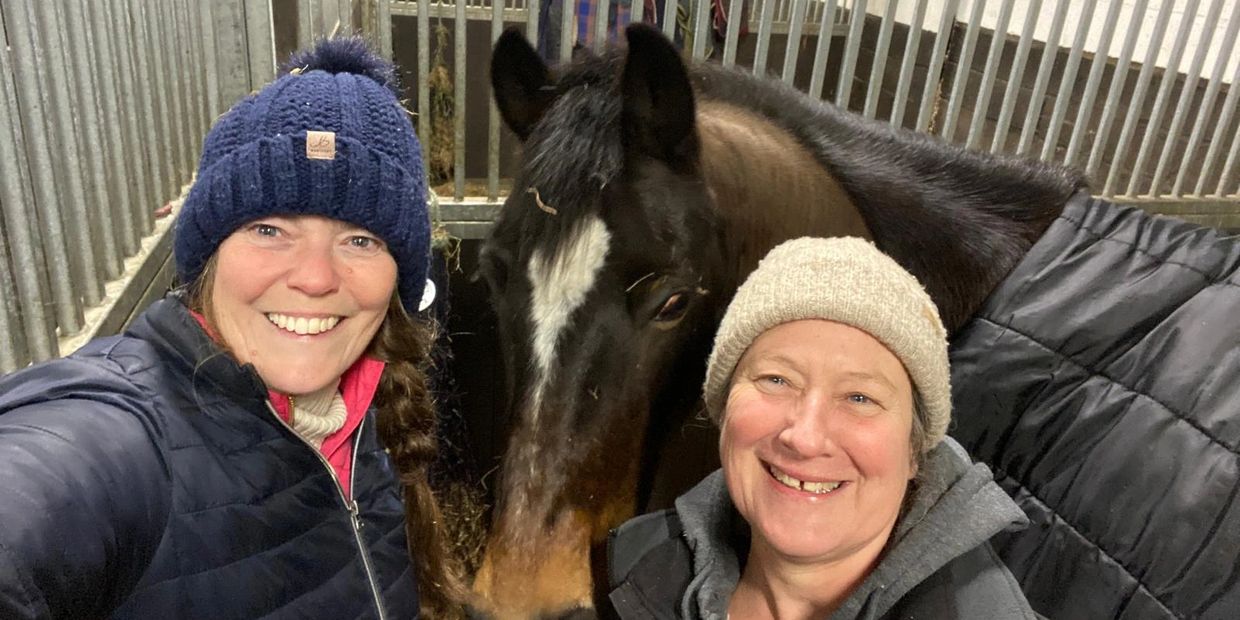Two women smiling with a horse in a stable, wearing warm hats and jackets.