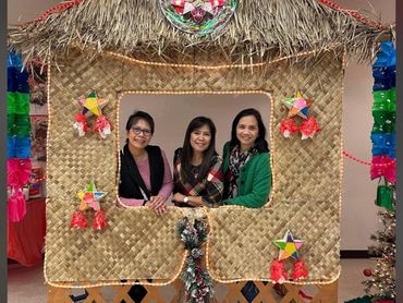 Three women posing inside a festive nipa hut decorated with star lanterns and poinsettias.