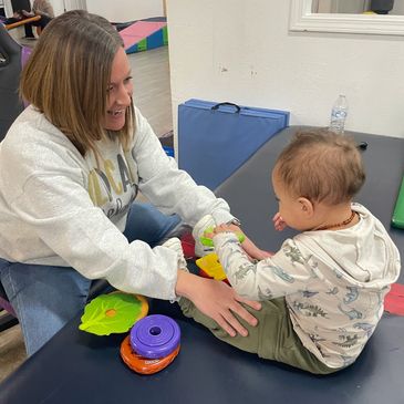 Woman engaging playfully with a child on a padded table with colorful toys.