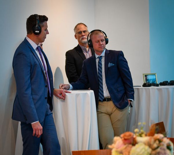 3 men standing at a table. 2 men are wearing headphones and appear to be listening to something.