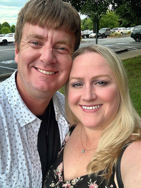 Smiling couple taking a selfie outdoors with trees and parked cars in the background.