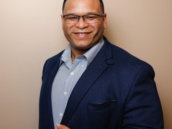 Confident man in glasses smiling, dressed in a navy blazer and striped shirt.