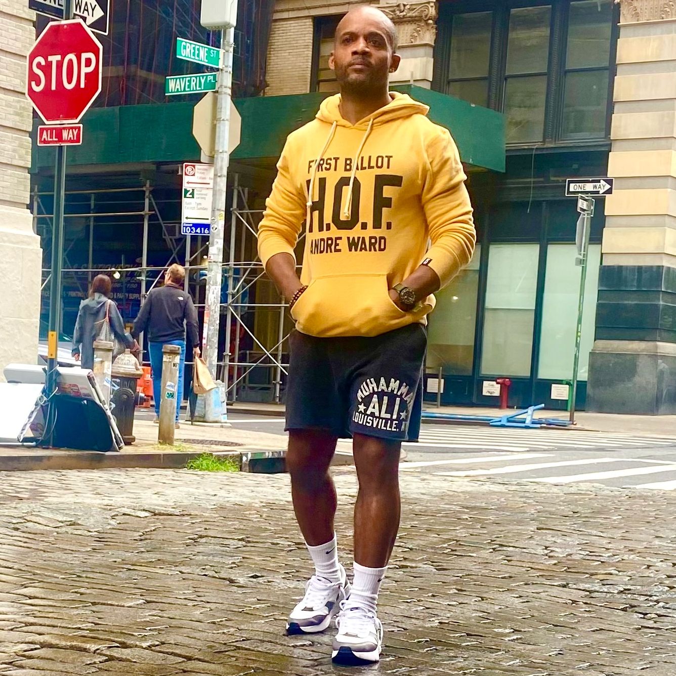 Man in yellow hoodie and black shorts standing on a cobblestone street.