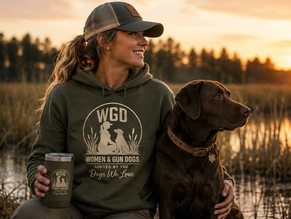Woman wearing a Women & Gun Dogs hoodie sitting beside a Labrador at sunset by a marsh.
