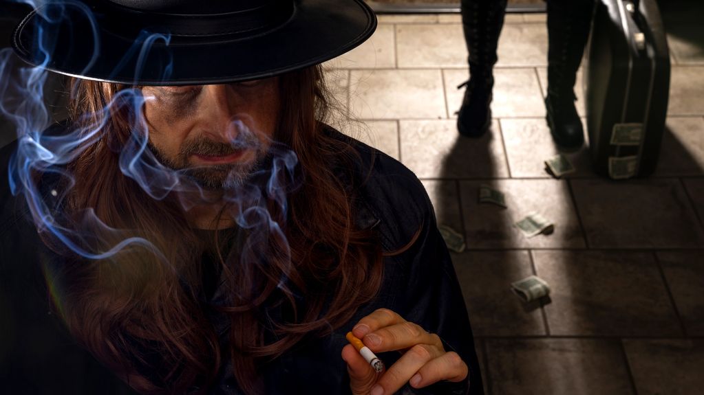 Man with a hat smoking a cigarette with money and a suitcase on the floor.
