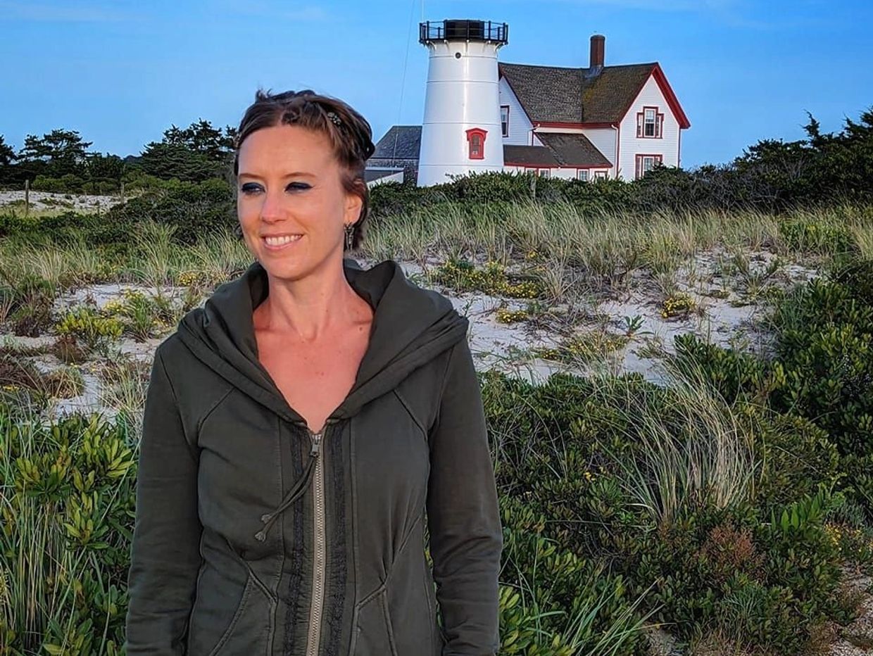 Woman smiling near a lighthouse surrounded by greenery under a clear blue sky.