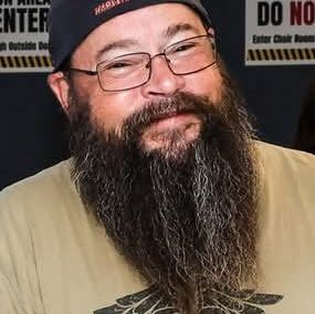 Smiling man with a long beard and glasses wearing a cap and T-shirt.