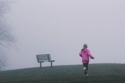 A girl wearing a pink jumper running up a grassed hill. The sky is dull. A black bench on the hill.