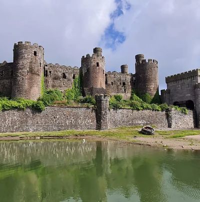 A medieval stone castle with towers by a calm moat under a partly cloudy sky.
