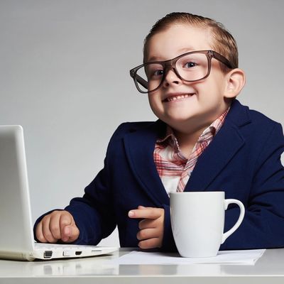 Young boy in glasses and suit sitting at laptop with coffee, smiling