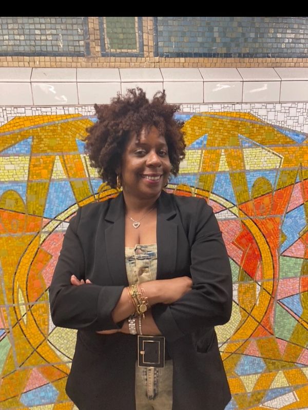 Smiling woman with curly hair stands confidently in front of colorful mosaic wall.