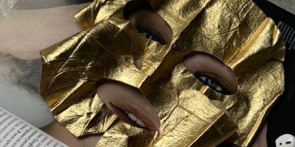 A gold facial mask with eye and mouth cutouts lies on a magazine page beside white earbuds.