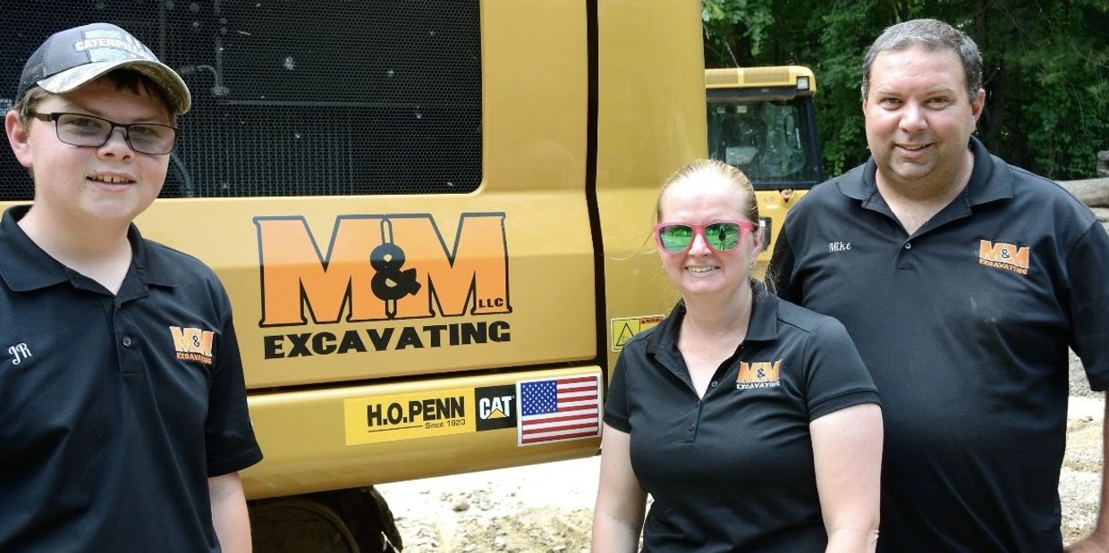 Three people in branded M&M Excavating shirts stand by heavy machinery outdoors.
