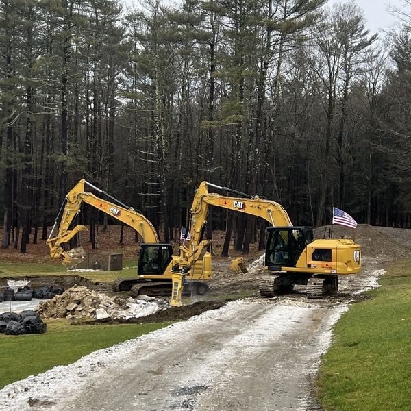 Two yellow CAT excavators working on a gravel road near a wooded area.