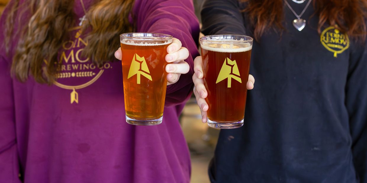Two people holding glasses of amber beer with brewery logos.