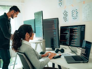 Two programmers work together at a desk with multiple monitors displaying code.