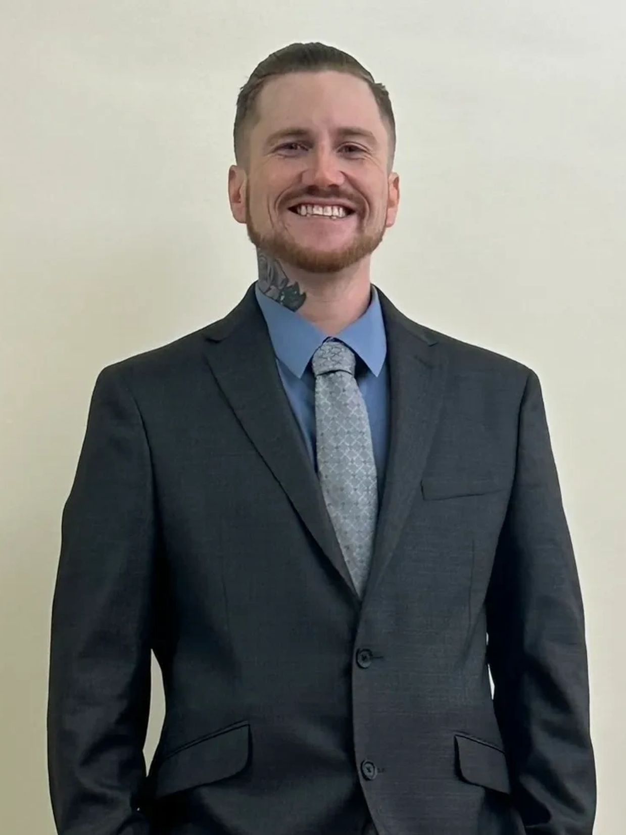 Smiling man in a dark suit and patterned tie with a neck tattoo.