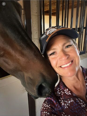 Smiling woman with a horse nuzzling her cheek inside a stable.