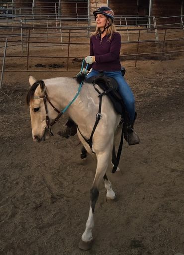 A woman in a helmet rides a cream-colored horse in an enclosed dirt arena.