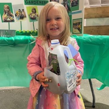 A young girl holding a milk jug with ladybugs, smiling at a gardening event.