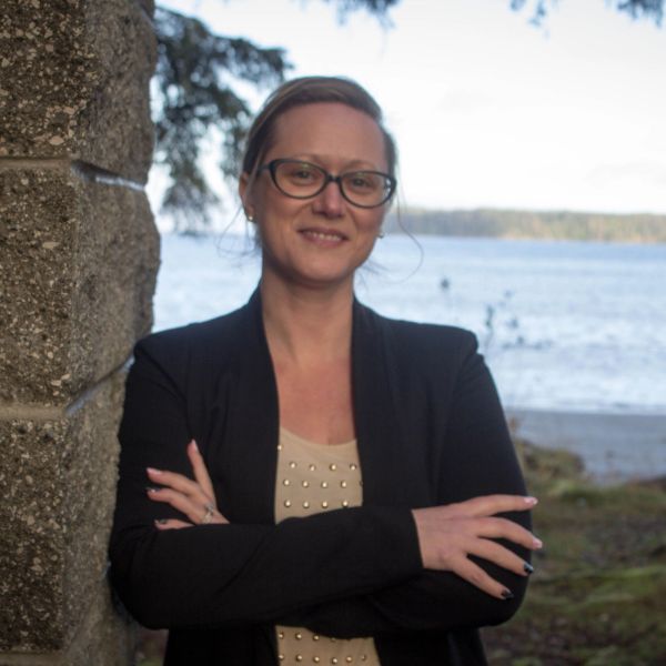 Woman with glasses leaning on a stone wall by the water.