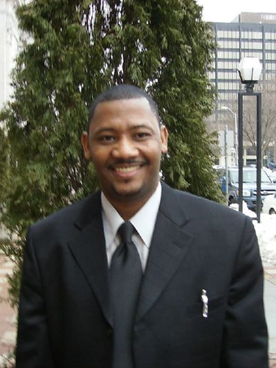 Man in a black suit smiling outdoors on a city sidewalk with snow around.