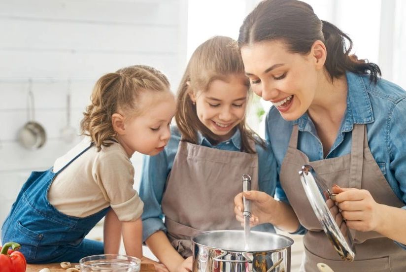 a mother cooking with her 2 daughters sitting on the countertop , all laughing and enjoying