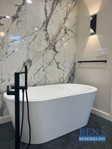 a stand alone tub, black finishing, white marble tiles with gold & grey veins by BEN's Remodeling