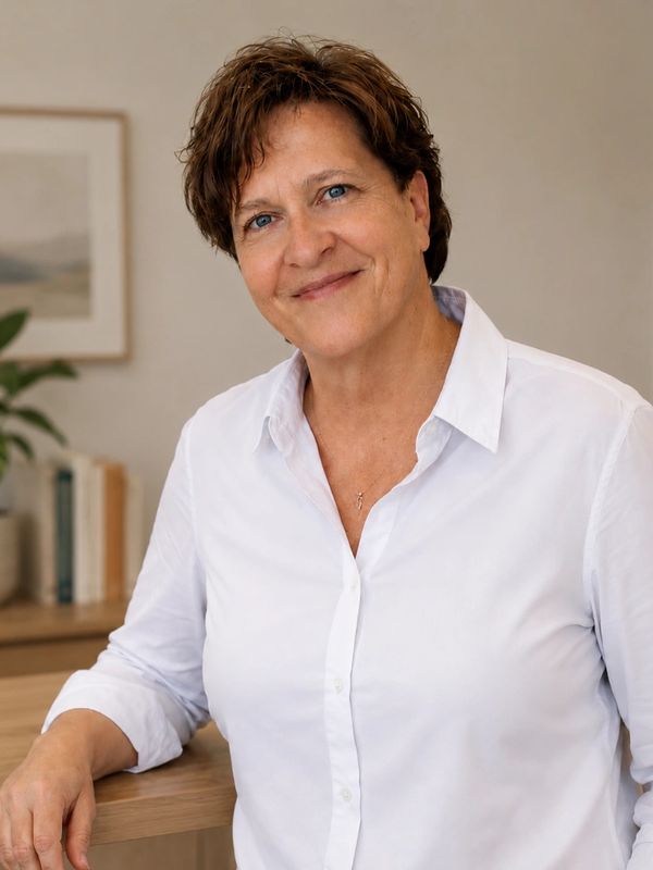 Smiling woman in a white shirt leaning on a wooden table in a cozy room.