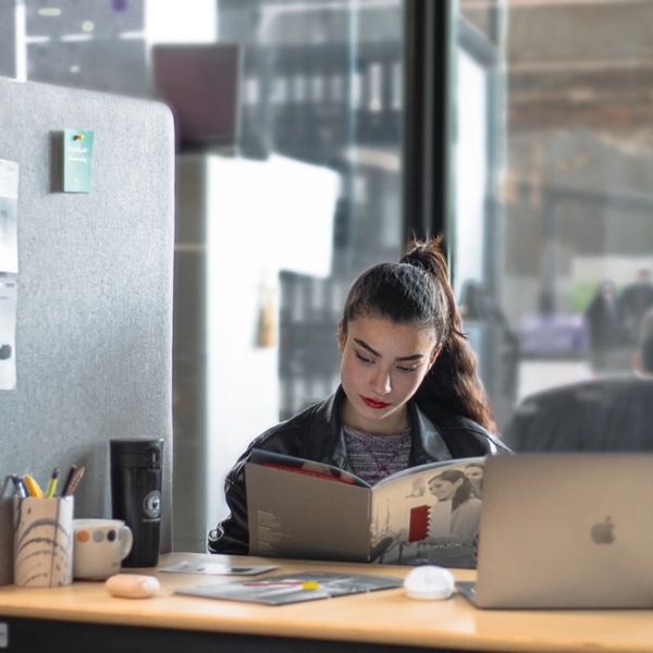 Young woman reading a magazine at a modern office desk with a laptop.