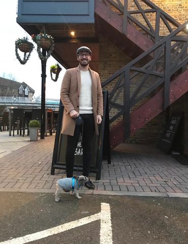 Man in a brown coat walking a small dog in a sweater outside a cafe.