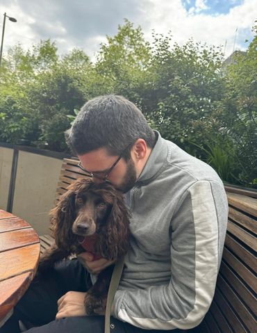 Man in gray jacket affectionately kisses his brown dog on the head while sitting on a wooden bench outdoors.