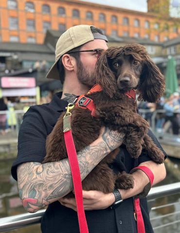 Man with tattoos holds a brown dog wearing a red harness.