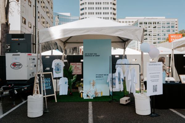 Outdoor market stall with shirts and gift boxes under a white tent.