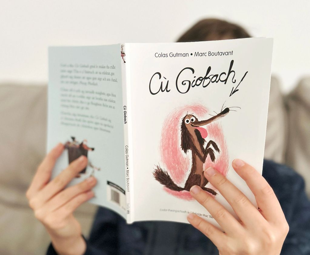 Child holding book reading the Gaelic language story of Cù Giobach/Scruffy Dog.
