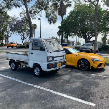 1986 Suzuki Carry by OIWA, juxtaposed with a yellow Toyota Supra, highlighting diverse vehicle offerings