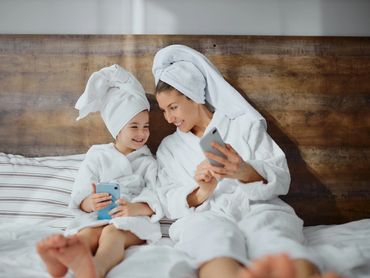 Mother and young daughter are in the bed with a towel on head and bathrobe on. Looking at mobile