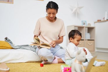 Mother is sitting on a mattress on the floor with young toddler nearby trying to figure things out
