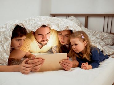 Parents & two children are in bed with cover over head like a tent while looking at computer.