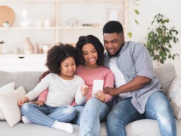 Family of 3 is watching a mobile device while sitting on the couch.