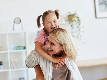 Mother has young daughter on her back. Young daughter is laughing and hugging the mom's neck