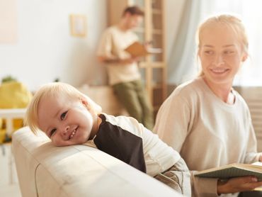 Mother watching her young toddler place head on back of couch as she reads to her. Dad is close