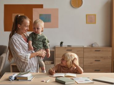 Mother is holding a toddler while her other daughter looks up at her.