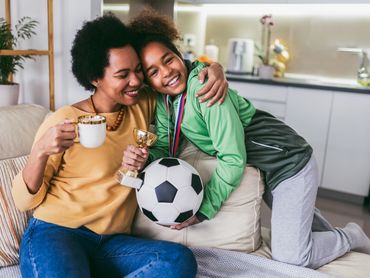 Mother is holding a cup of coffee in her hand while hugging daughter who has a soccer ball in hand