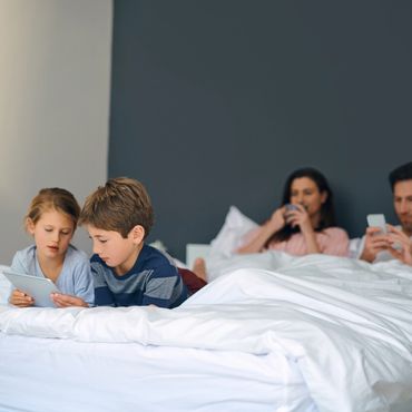 Parents lying at the top of the bed. Their 2 children are lying at the bottom of bed with device