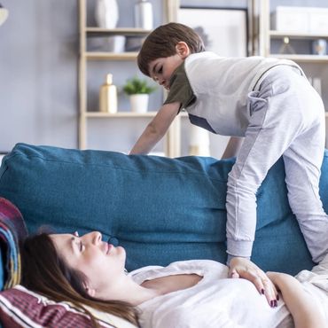 Little son jumps on the couch and leans on the back of the sofa where his mom is resting.
