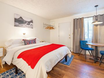 Hotel room with bed and red blanket, blue throw rug and small table with blue chairs