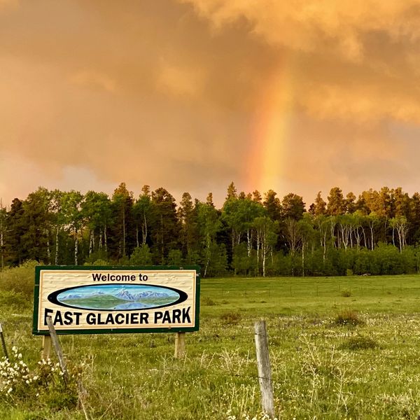 A colorful sign saying Welcome to East Glacier in front of a forest and stormy sky with a rainbow.